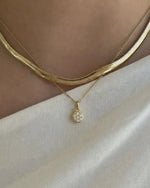 A close-up of a person's neck and upper chest wearing layered gold necklaces: a thicker snake-style chain and a thinner chain with a small round pavé pendant, paired with a white top.