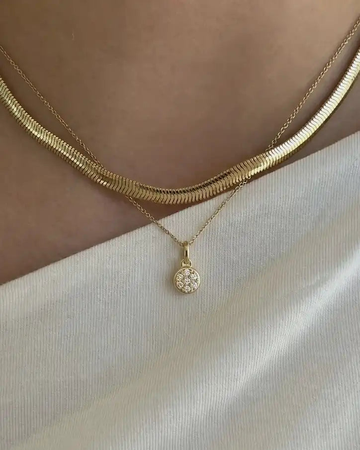 A close-up of a person's neck and upper chest wearing layered gold necklaces: a thicker snake-style chain and a thinner chain with a small round pavé pendant, paired with a white top.