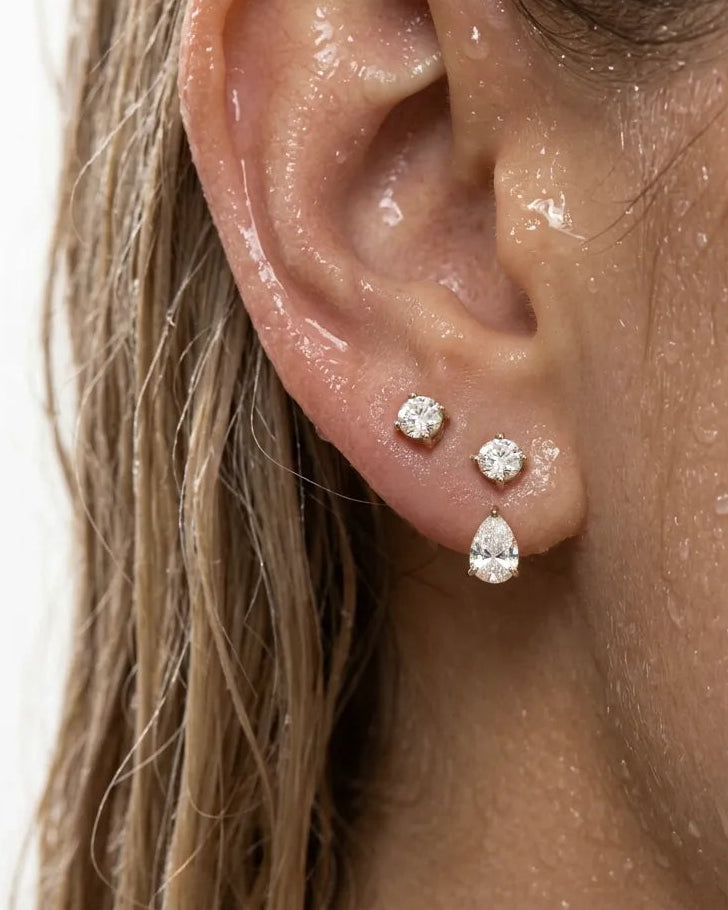 Close-up of a person's ear wearing three stud earrings: two small round diamonds and one larger pear-shaped diamond, shown against skin and hair with water droplets.