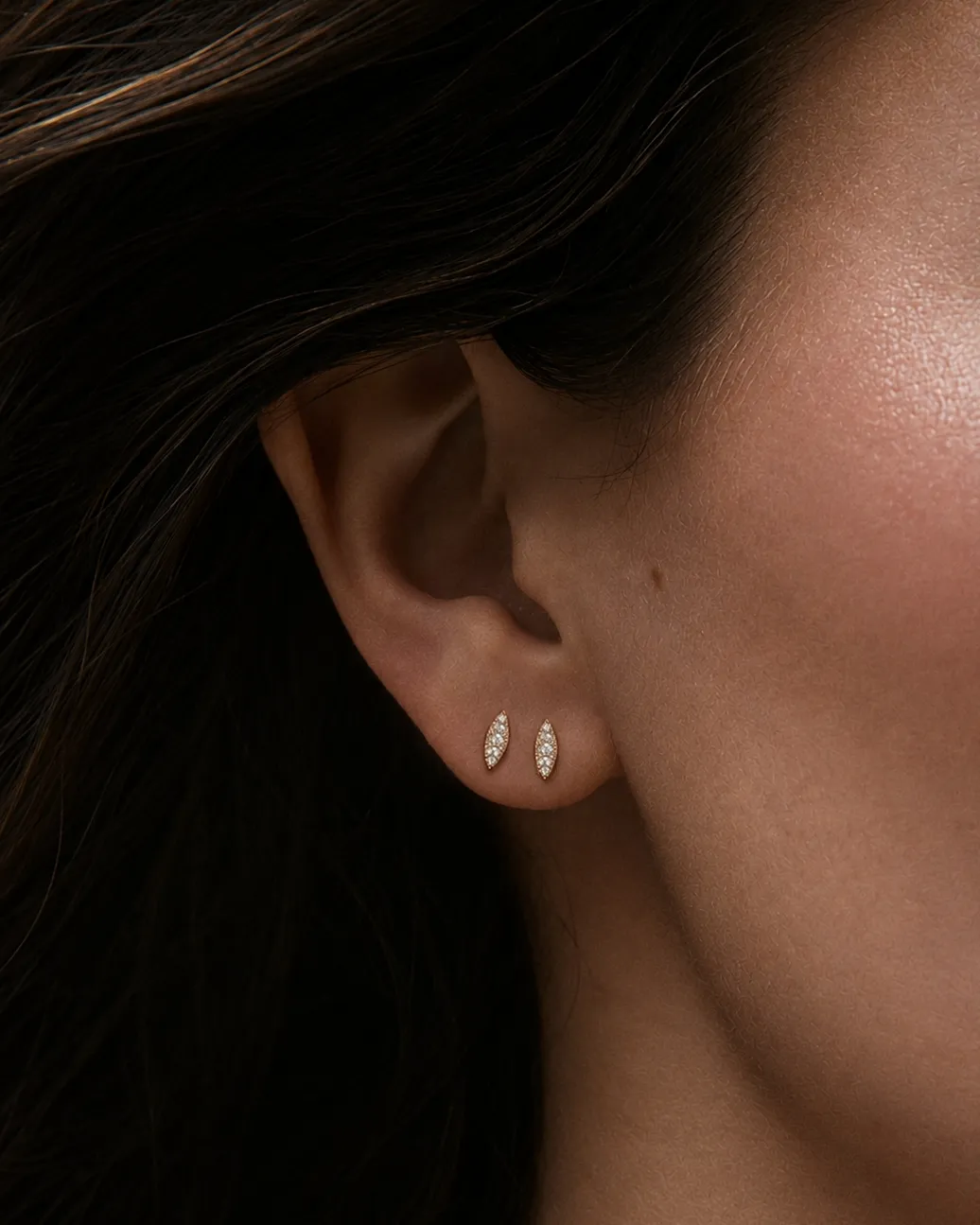 Close-up of a woman's ear wearing a pair of small gold leaf-shaped stud earrings with clear stones, set against dark hair and soft warm lighting.