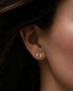Close-up of a woman's ear wearing a pair of small gold leaf-shaped stud earrings with clear stones, set against dark hair and soft warm lighting.