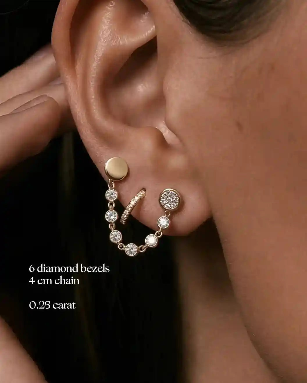 A close-up of a model's ear wearing a gold huggie hoop earring in the 2nd stud adorned with six round-cut diamond bezel  in a 4 cm chain from the first and third piercing alongside a small gold disc stud and a diamond-accented disc stud on respective earrings. 